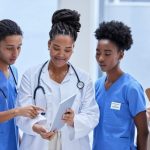 A group of ethnically diverse female healthcare workers, including a doctor and nurses, are gathered together looking at a digital tablet, discussing patient care in a hospital setting.