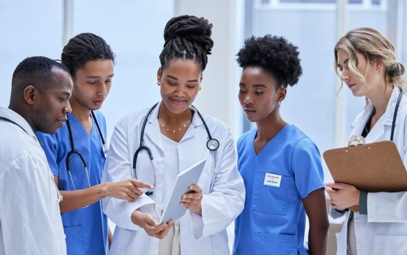 A group of ethnically diverse female healthcare workers, including a doctor and nurses, are gathered together looking at a digital tablet, discussing patient care in a hospital setting.